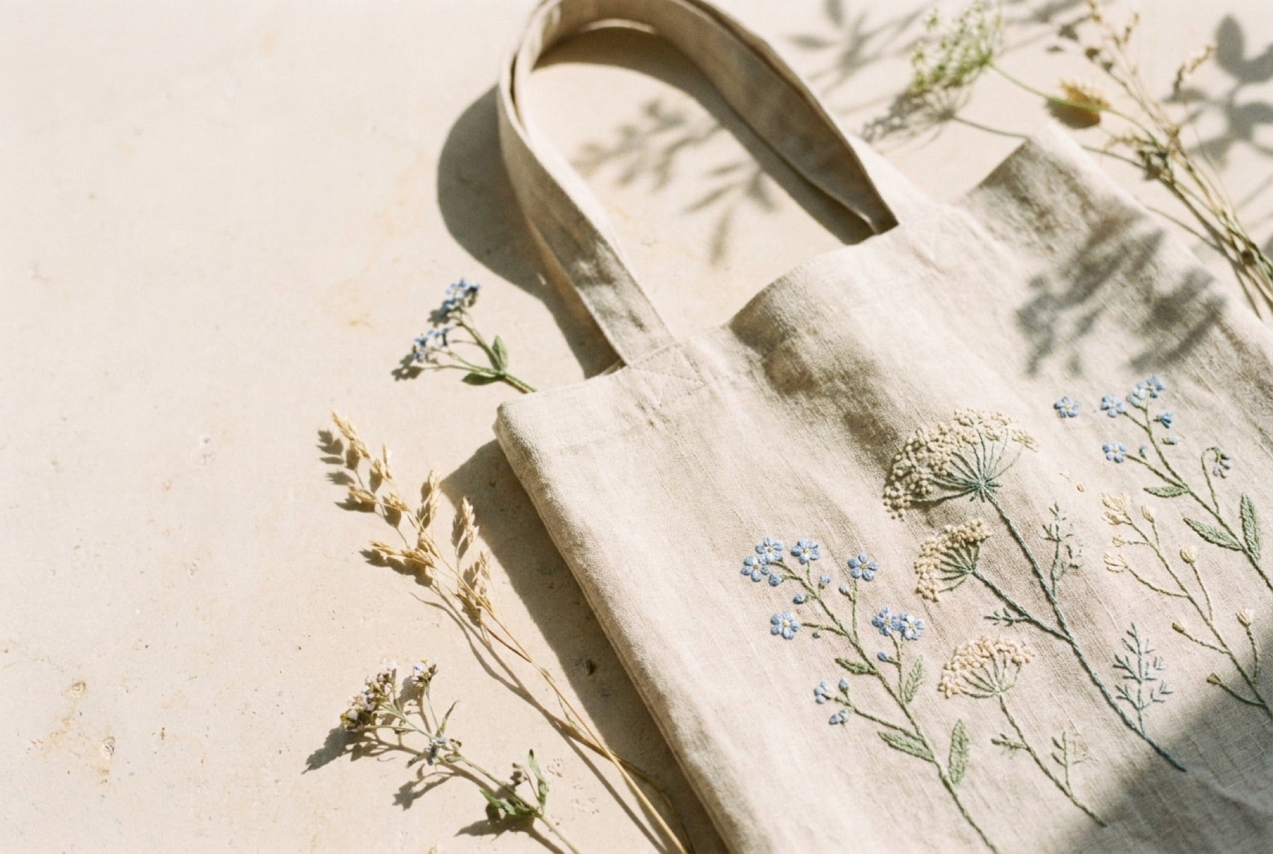 Natural linen tote bag embroidered with blue wildflowers, resting in warm sunlight